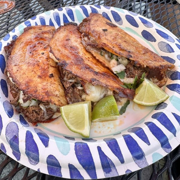 Three birria tacos with melted cheese and shredded beef, garnished with lime wedges on a paper plate.